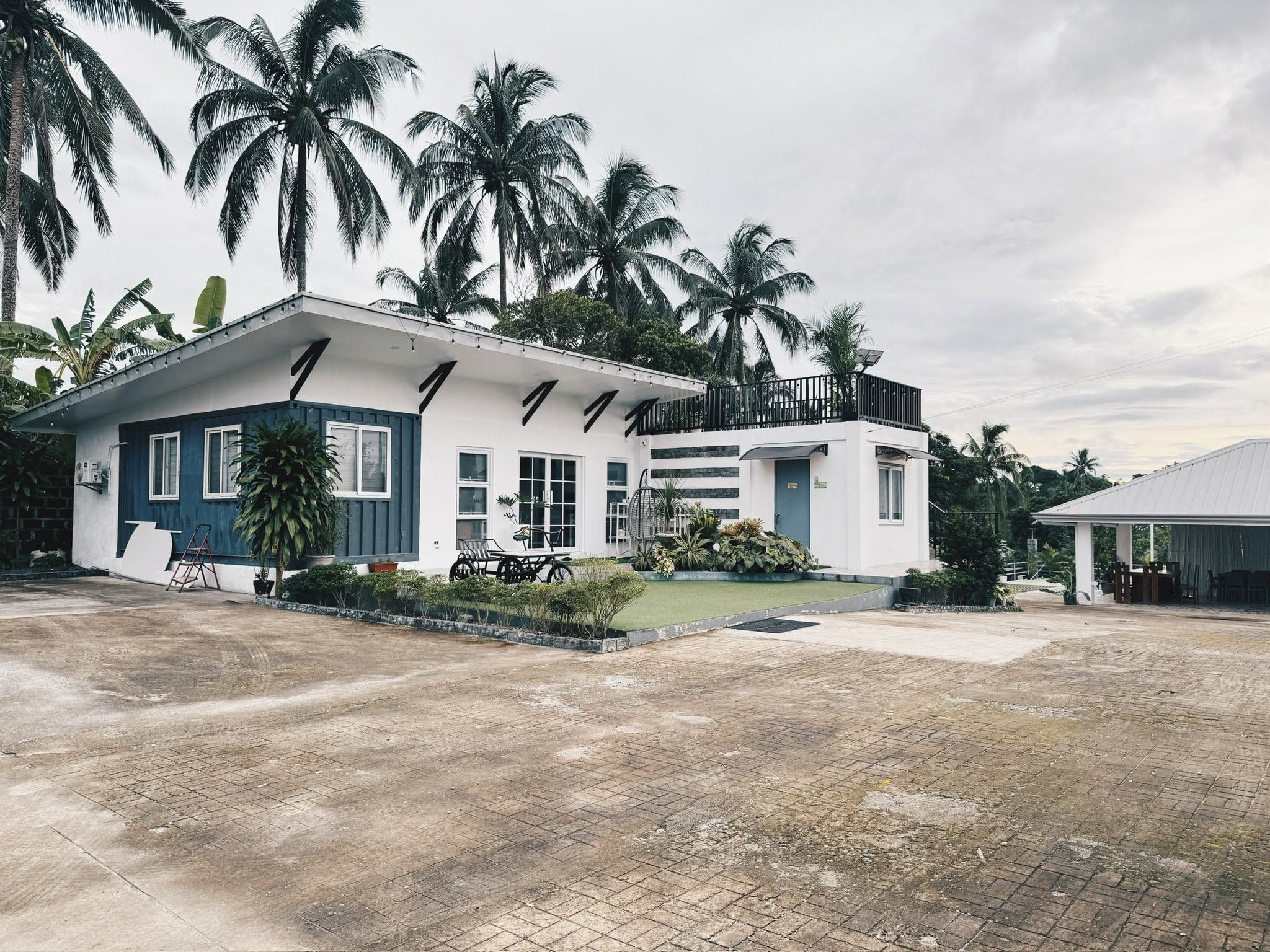 The white main house with navy board walls and a roof deck, seen across the paved parking area under coconut palms