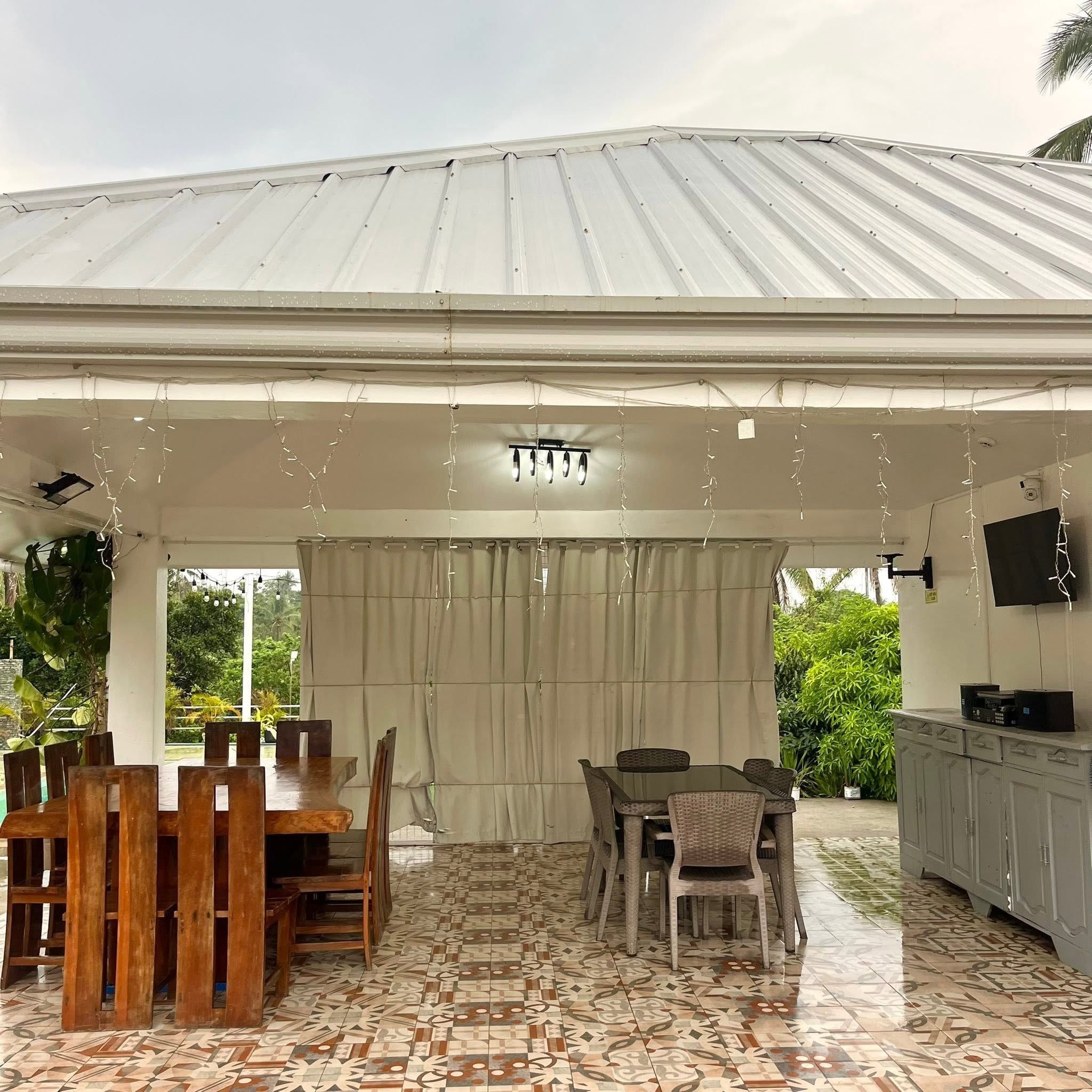 The open-sided pavilion with a long wooden dining table, a second dining set, a TV and a sideboard on patterned tiles