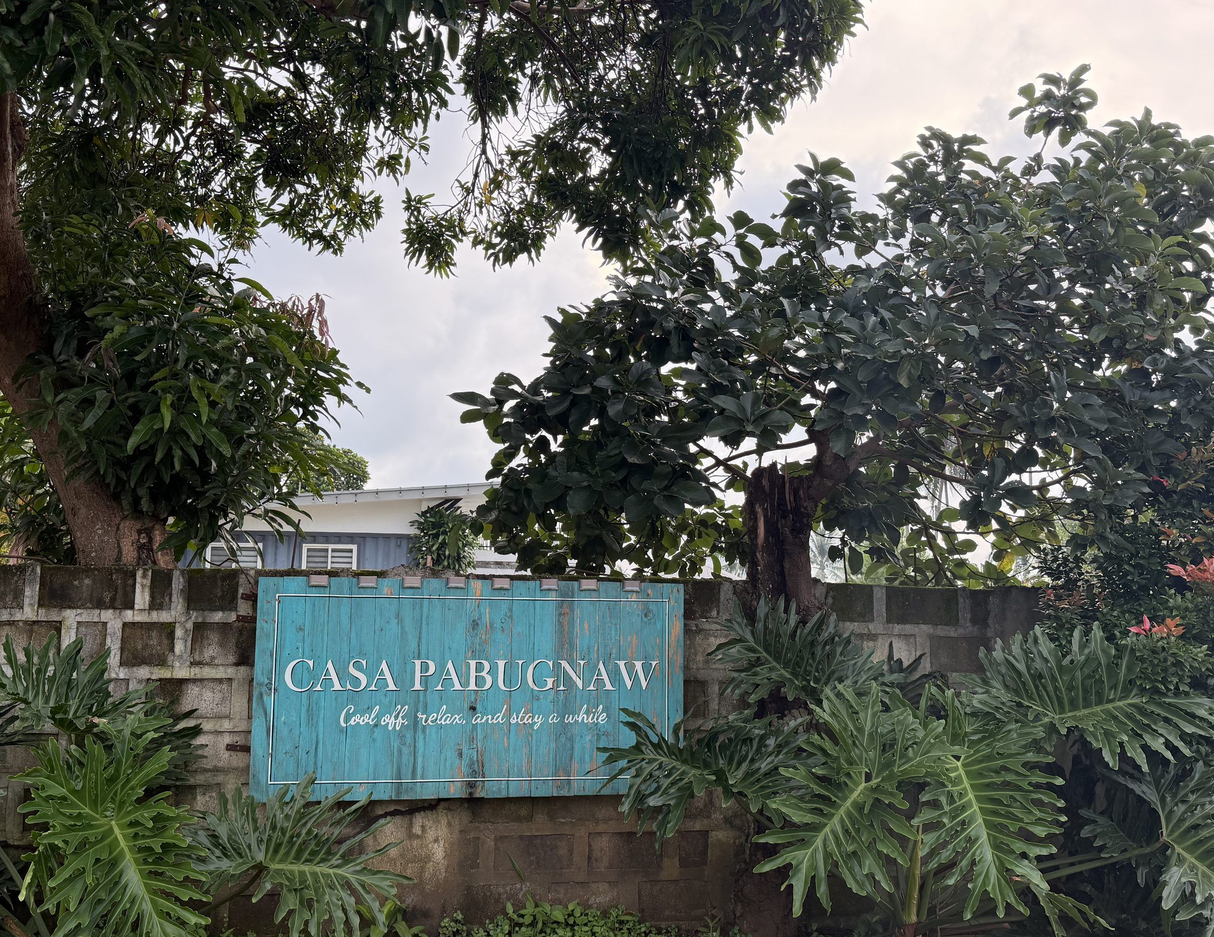 The teal Casa Pabugnaw sign on the front wall among monstera and shade trees