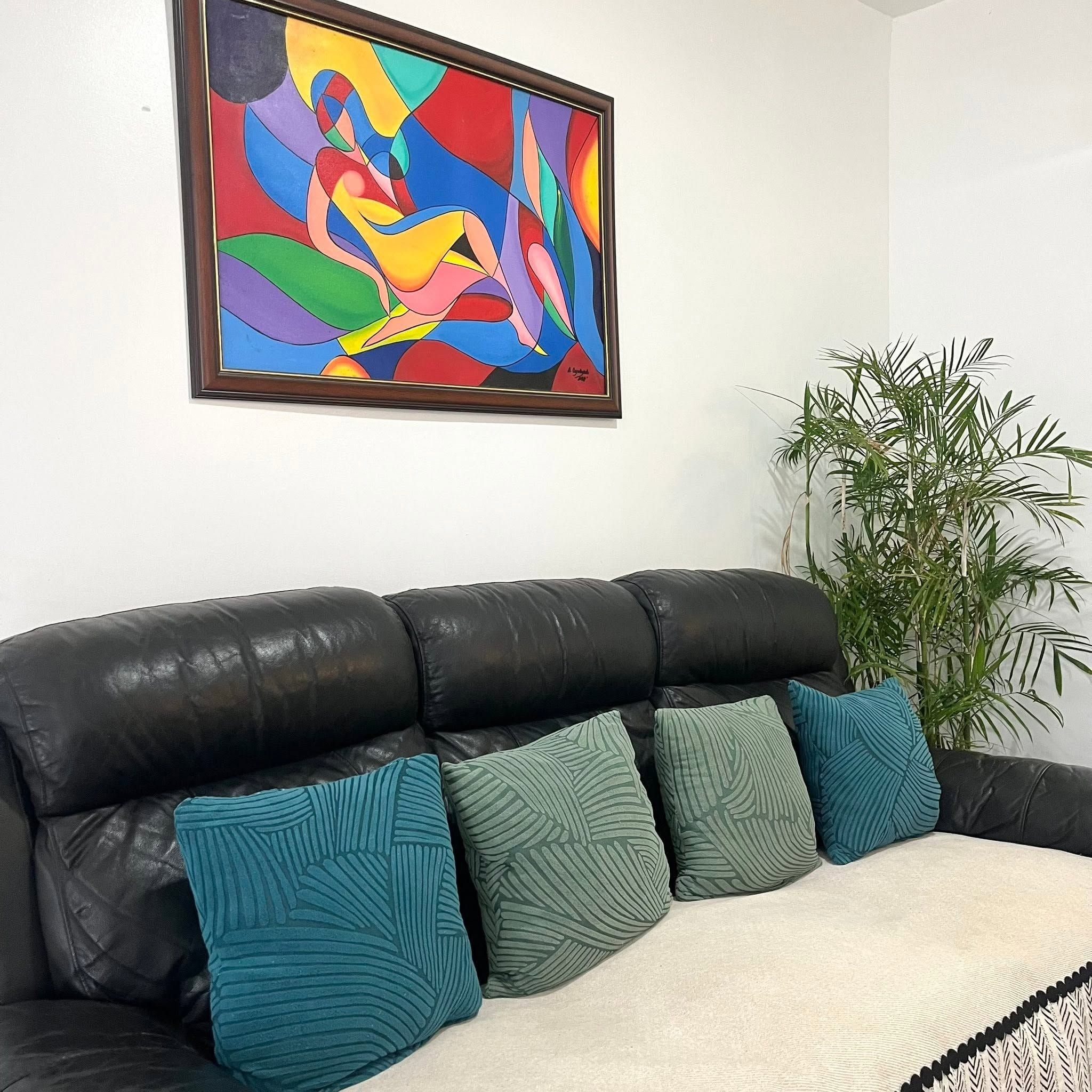 Black leather sofa with teal and green cushions below a colourful abstract painting, beside a potted palm